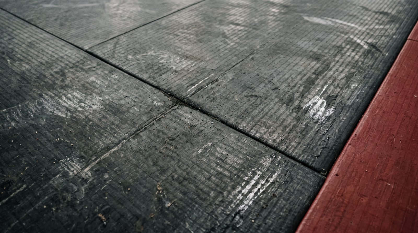 Tatami noir usé, voilé d’un film gris, poussière tassée dans le grain et dans le joint entre deux dalles